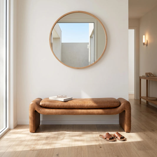 A modern curved leather bench in a warm tan-brown color, styled in a minimalist entryway with a round wooden-framed mirror on the wall, light wood flooring, and a pair of brown sandals on the floor.