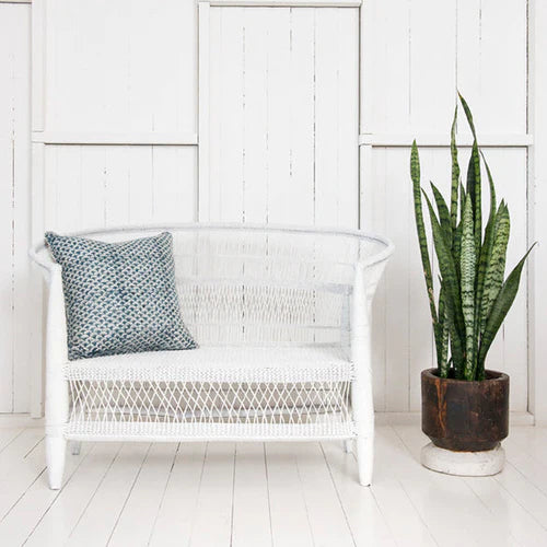 White wicker chair with a patterned pillow and a potted plant in a room with white walls and floor.
