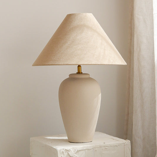 Beige table lamp with a textured shade on a marble surface against a neutral wall.