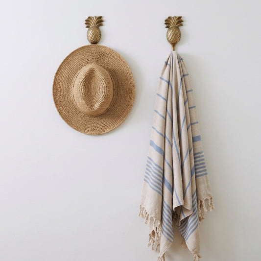 A woven straw sun hat hangs from a pineapple-shaped wall hook on a textured white wall, next to a beige and blue striped fringed scarf hanging from a matching hook.