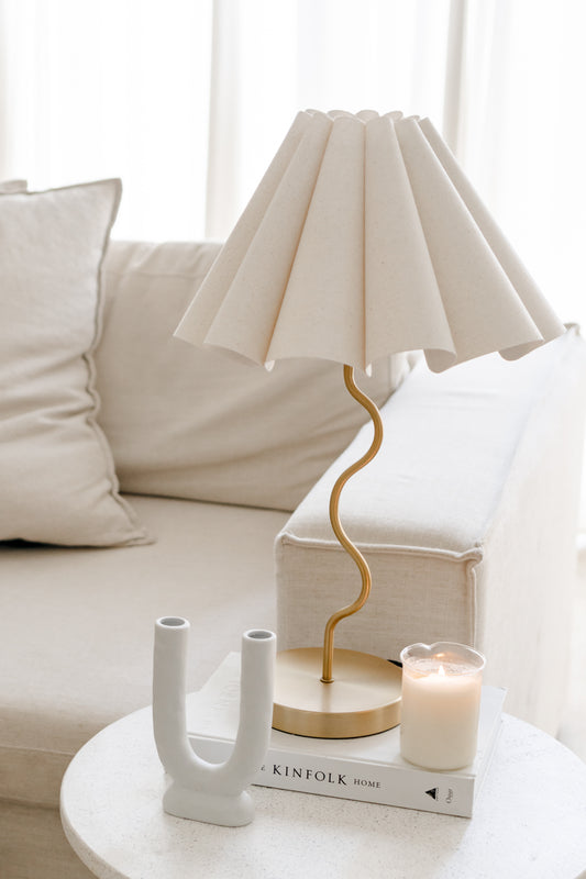 White lamp with gold base on a table next to a candle and decorative book in a living room setting.