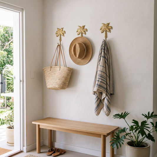 A styled entryway corner with a light wood bench against a white wall, a woven straw tote bag hanging on the left hook, a straw hat hanging in the center, and a striped towel hanging on the right. Three gold palm-shaped wall hooks are mounted above the bench, with a glass door and potted plants visible.