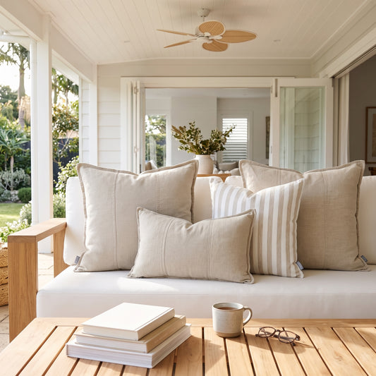 A set of neutral-toned throw pillows, including solid beige and cream-and-white striped pillows, arranged on a white outdoor sofa on a covered porch with a wooden slat coffee table in the foreground.