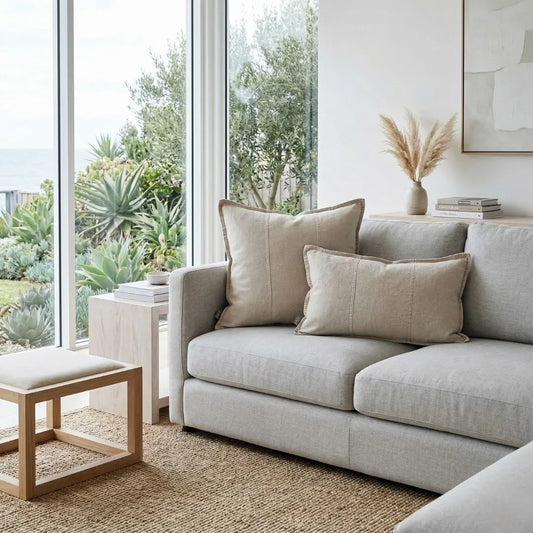 Light gray sofa with beige pillows in a modern living room with large windows and plants.