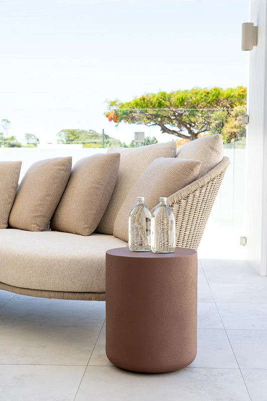 Beige outdoor sofa with a woven backrest and a small round brown table with decorative bottles on a patio.