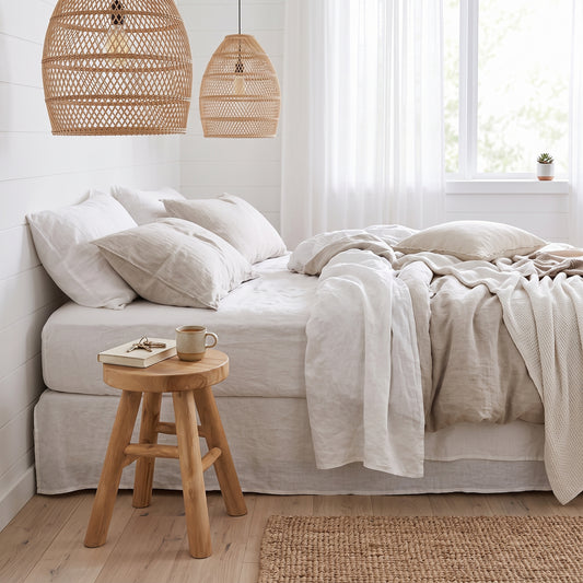 A neatly made bed with white and beige linens in a bright bedroom, with two woven pendant lights hanging above, a small wooden stool with a mug, and a window with sheer curtains in the background.