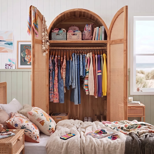 A large arched wooden wardrobe or closet with cane-style doors stands open in a bright bedroom, filled with hanging clothes and storage baskets. A bed with patterned pillows and rumpled cream bedding is in front, and a window with a beach view is on the right.