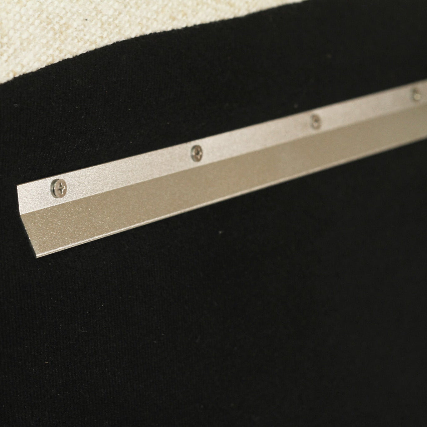 Close-up of a metallic silver metal track or bar with screws, mounted against a black textured background with a cream fabric corner visible at the top left.