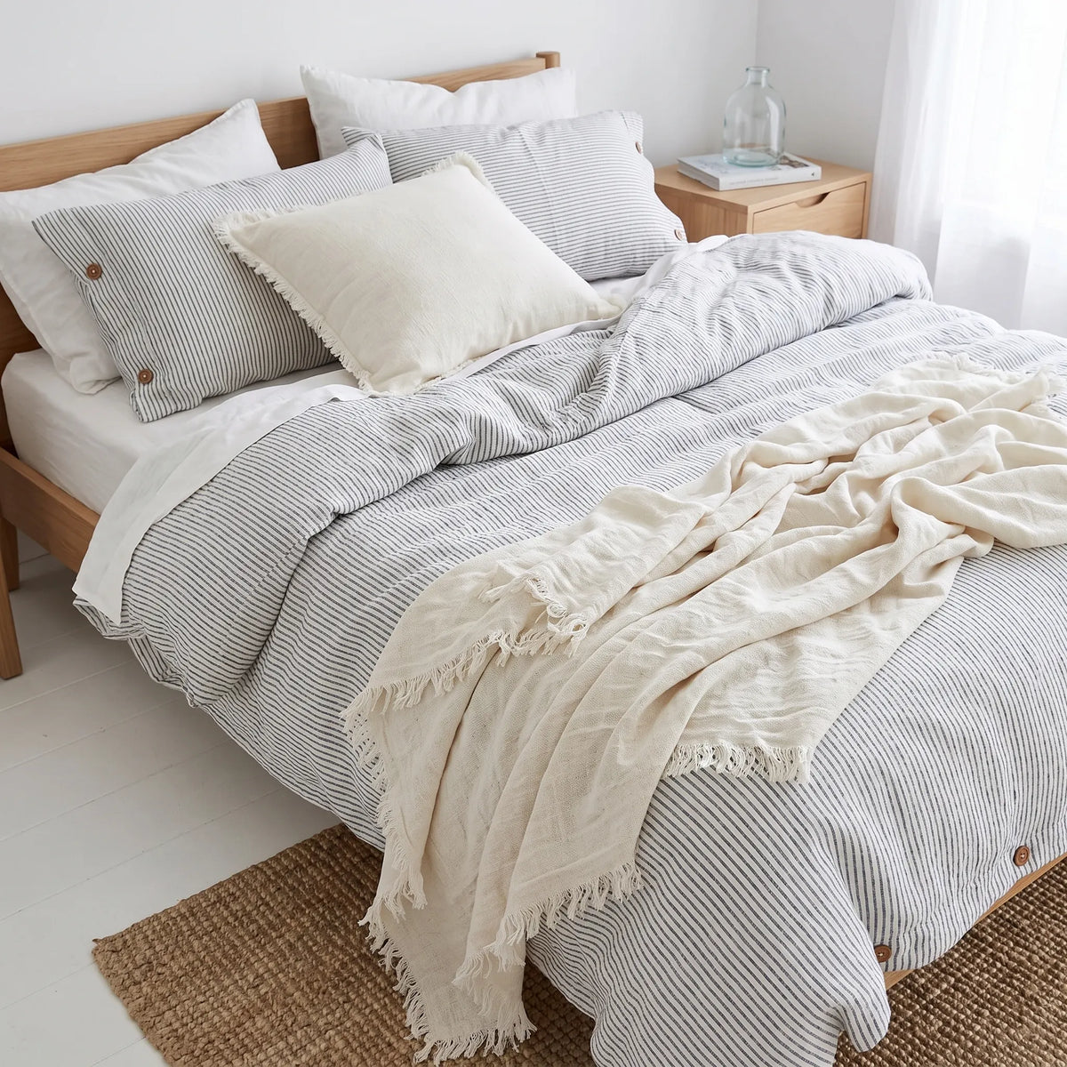 A neatly made bed in a bright bedroom setting, styled with a striped bedding set in gray and white, a cream pillow sham, and a cream throw blanket with fringe.