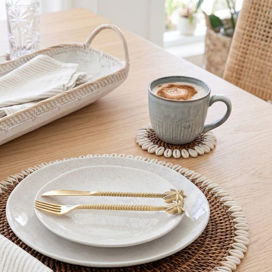 Café setting with a cup of coffee, white plate with gold cutlery, and woven placemat on a wooden table.
