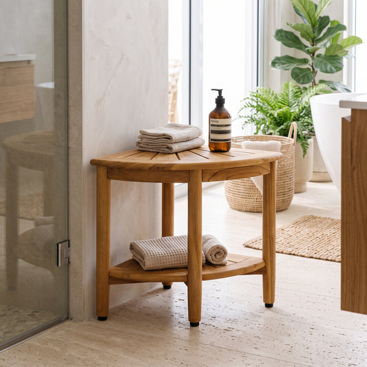 A small round wooden corner stool with a lower shelf, styled in a bright, modern bathroom next to a glass shower door. The stool is decorated with folded beige towels and an amber bottle of soap, with a wicker basket and potted plants in the background.