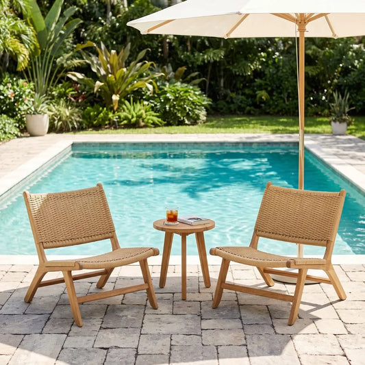 Two woven rattan lounge chairs with a small round side table and a cream patio umbrella, arranged on a stone patio next to a swimming pool with lush green garden foliage in the background.