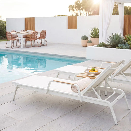 White outdoor patio chaise lounge chairs with light wood armrests arranged beside a swimming pool on a stone patio, with a dining set and plants in the background.