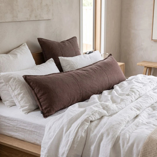 A bedroom scene showing a bed with white bedding and multiple brown and white pillows, including a long brown lumbar pillow in the foreground.