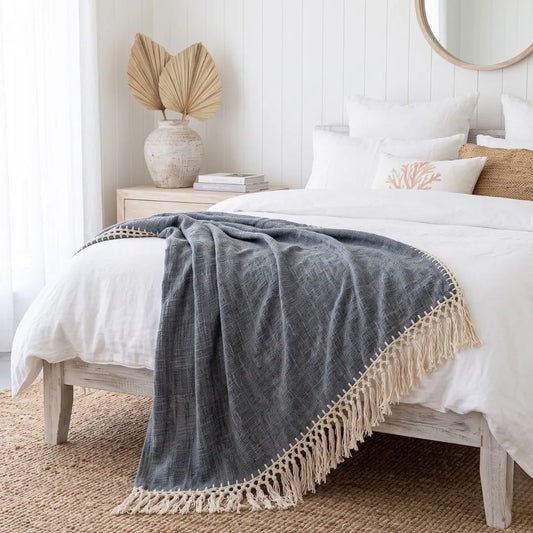 A dark gray textured throw blanket with cream fringe trim draped over a white bed in a bright, modern bedroom setting.