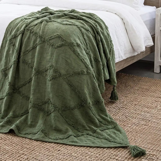 An olive green textured throw blanket with a raised geometric diamond pattern and tassels, draped over a bed with white linens on a woven jute rug.