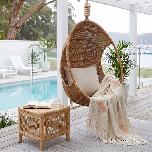 A woven hanging egg chair with a cream cushion sits on a wooden deck beside a small rattan side table with an open book and a cream throw blanket featuring coral palm tree prints. A swimming pool, white lounge chairs, and potted plants are visible in the background.