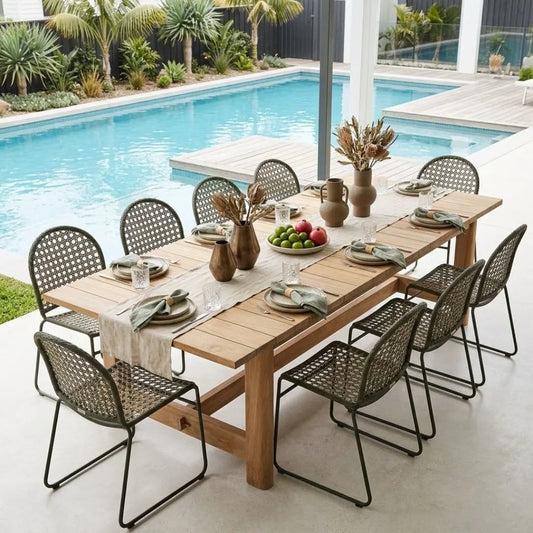 A wooden dining table set with multiple woven metal chairs, styled with tableware and decorative vases on a patio overlooking a swimming pool.