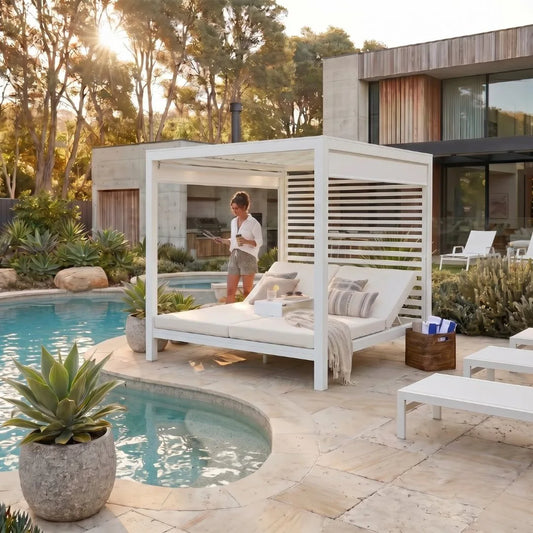 A white outdoor cabana-style lounge set with a chaise daybed, cushions, and a throw blanket, situated beside a swimming pool on a stone patio. A person is standing under the cabana, and a modern house and trees are in the background.