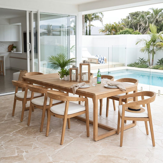 A modern wooden dining table and six matching chairs with white upholstered seats, arranged on a stone patio overlooking a poolside outdoor living area.