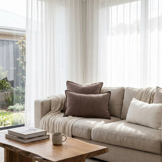Beige sofa with brown and white cushions in a bright living room with large windows.