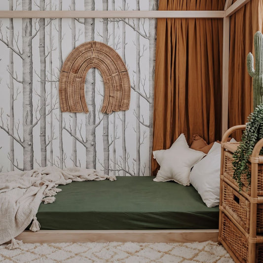 A cozy daybed styled in a bohemian bedroom setting, featuring a dark green fitted sheet, white and brown pillows, a beige throw blanket, a wooden canopy frame, and a distinctive birch tree wallpaper with a woven wicker arch decoration.