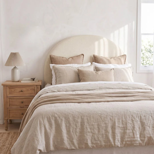 A neatly made bed in a softly lit bedroom, dressed in neutral beige and cream bedding with matching pillows. A light wood nightstand with a pleated lamp sits beside the bed against a white wall.