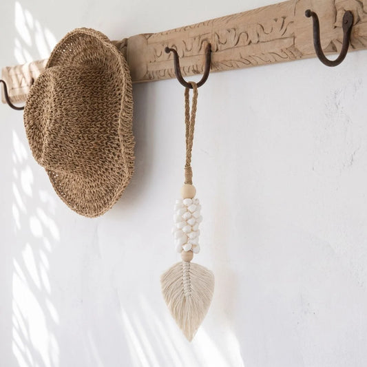 A woven straw hat hangs on a carved wooden wall hook with dark metal S-hooks, alongside a cream macrame tassel ornament.