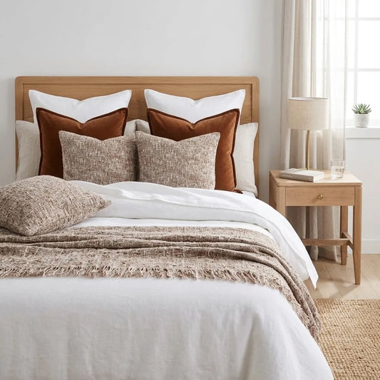 A neatly made bed in a bright bedroom setting, featuring white linens, a beige textured throw blanket, rust brown velvet and linen pillows, and a light wood headboard and nightstand.