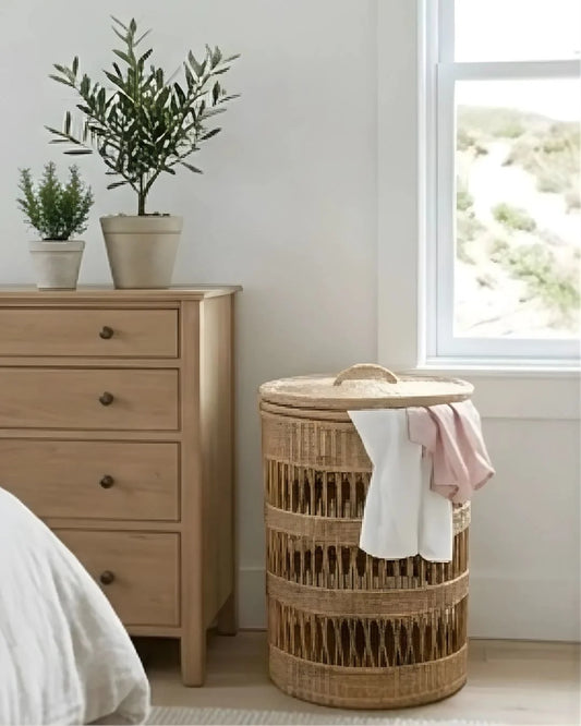 Woven Cane laundry basket with clothes next to a wooden dresser in a room with a window.