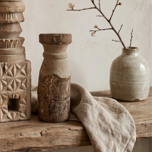 Wooden candle holder with textured surface on a wooden shelf with a vase and branch in the background.