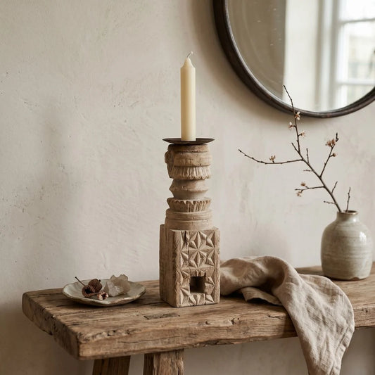 Wooden candle holder with a candle on a rustic wooden table, with a vase and branch in the background.