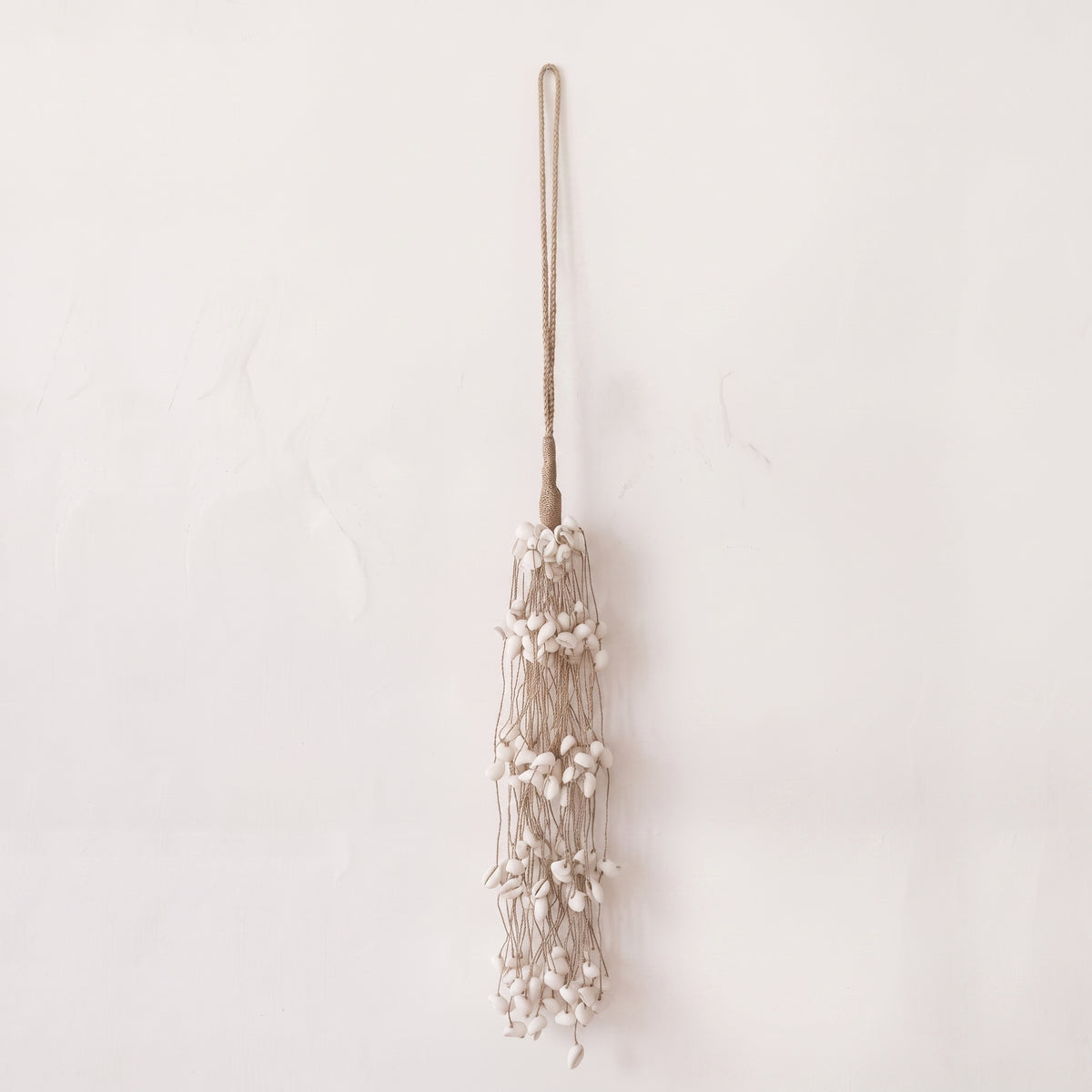 A bohemian-style tassel wall hanging made of beige cord, small white beads, and dried flower elements, suspended against a plain off-white wall.