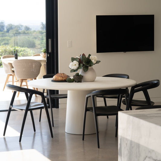 Modern dining area with a white round table and black chairs, featuring a TV on the wall. Featuring the Elio Round Microcement Dining Table – Seashell Finish from Sun Republic
