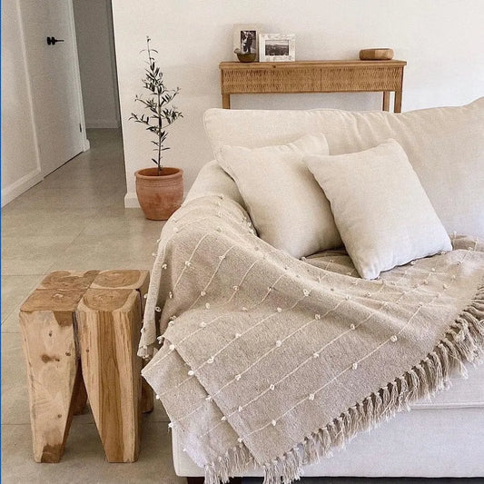 Beige textured blanket draped over a white sofa with teak peg stool and a plant in the background.