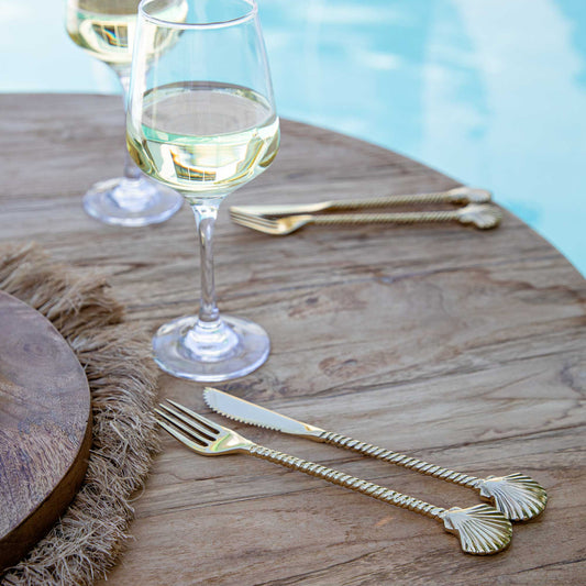 A set of gold brass cutlery with clam shell designs, including a knife and a fork, on a wooden table with a glass of wine in the background.