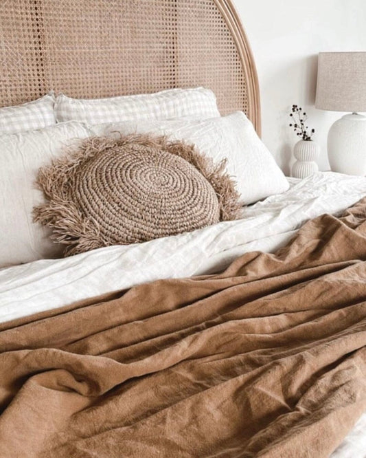 A round, natural raffia cushion with fringe, in a brown color, on a bed with a beige pillow, in a room with a wicker bed frame and a white lamp.