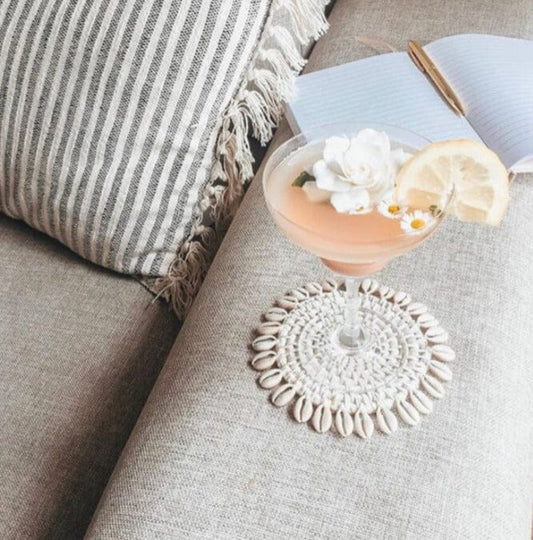 A round, white washed rattan coaster with a cowrie shell border, displayed on a sofa next to a cup with a lemon slice and a straw.
