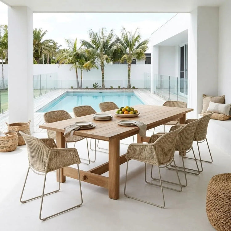 A modern outdoor dining set featuring a rectangular light oak wood table and eight woven wicker chairs with chrome metal legs, styled on a white patio with a swimming pool and palm trees in the background.