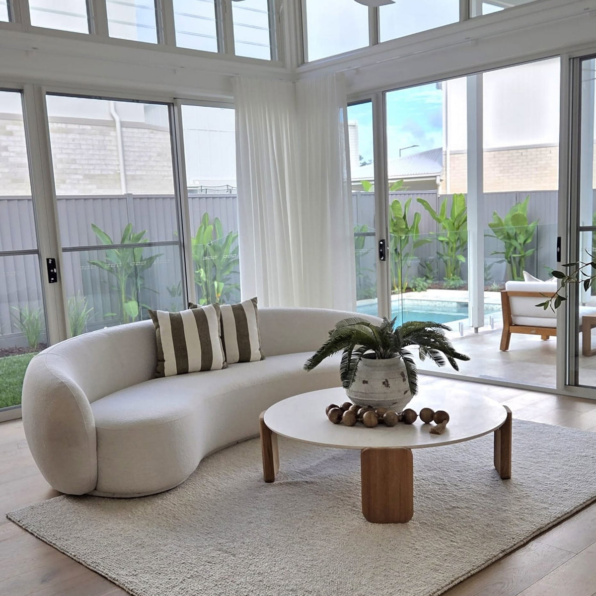 A modern curved cream sofa in a bright, sunlit living room with large windows, a round wooden coffee table, and a light rug.