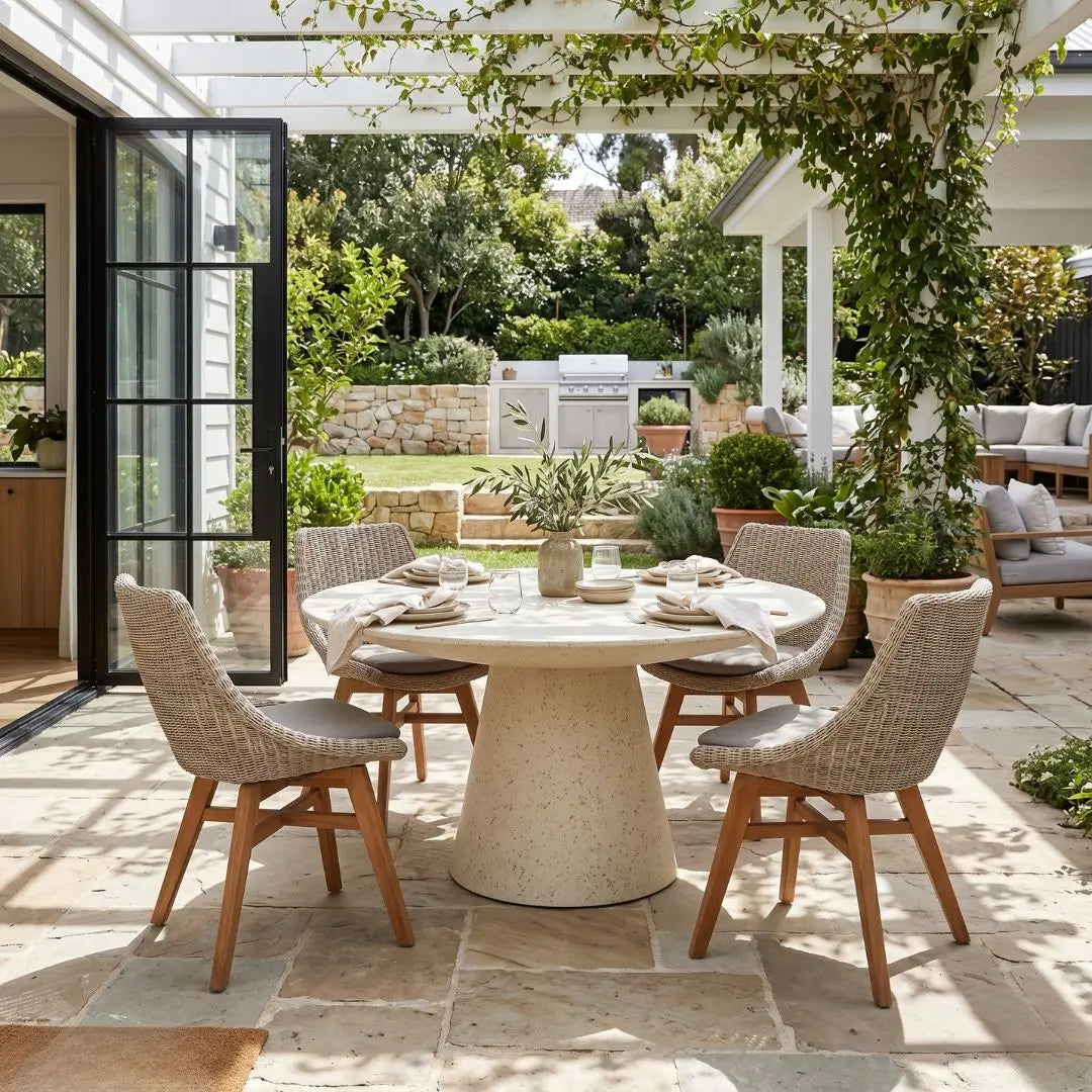 Outdoor patio with a round stone table and wicker chairs, surrounded by greenery.