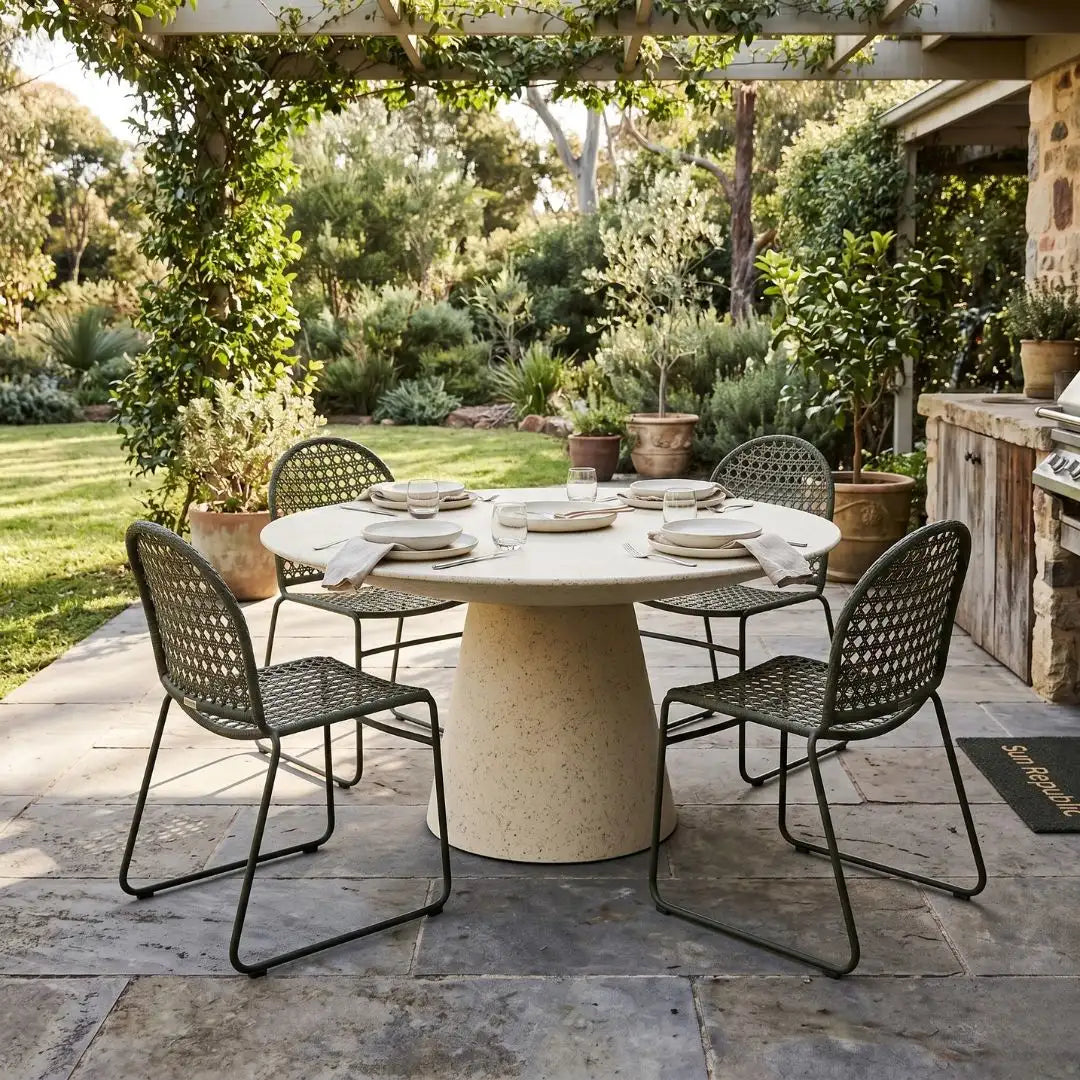 Outdoor dining setup with a round table and chairs on a stone patio, surrounded by greenery.