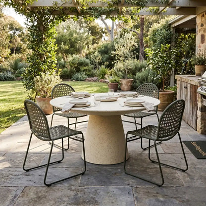 Outdoor dining setup with a round table and chairs on a stone patio, surrounded by greenery.