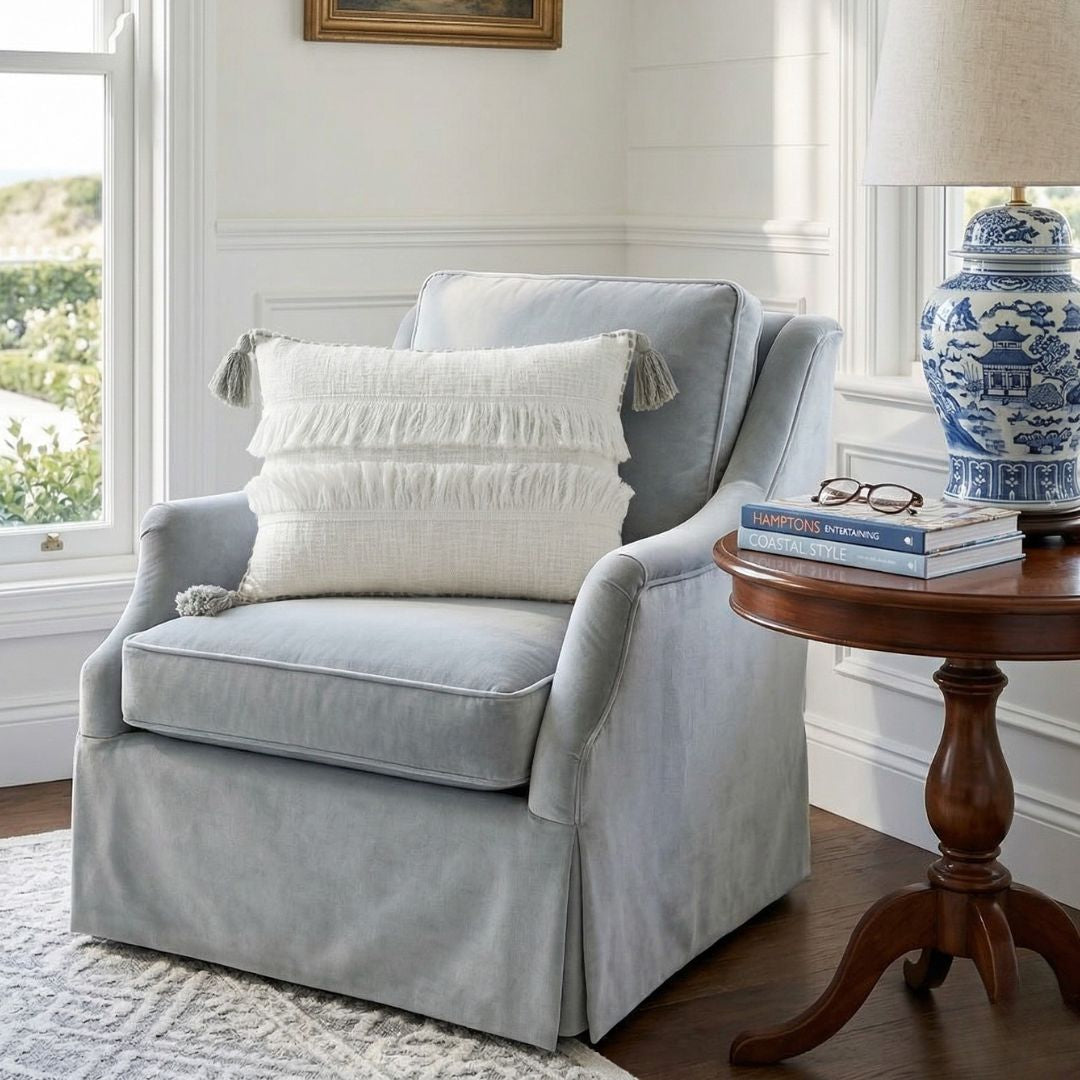 Gray armchair with a white pillow and wooden side table in a room with large windows.