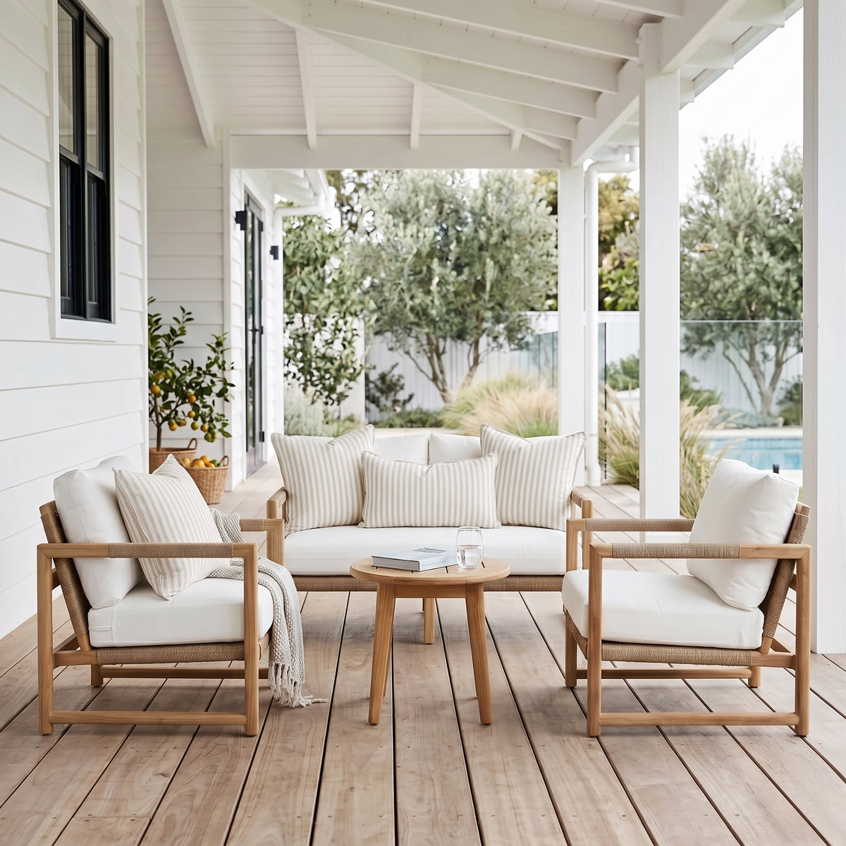 A 4-piece outdoor patio conversation set on a wooden deck under a white porch. The set includes a light wood frame, cream cushions, a matching 3-seater sofa, two armchairs, and a round wooden coffee table.
