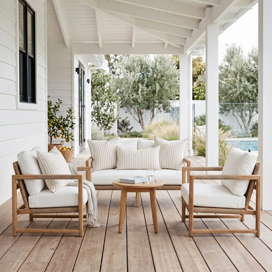 A 4-piece outdoor patio conversation set on a wooden deck under a white porch. The set includes a light wood frame, cream cushions, a matching 3-seater sofa, two armchairs, and a round wooden coffee table.