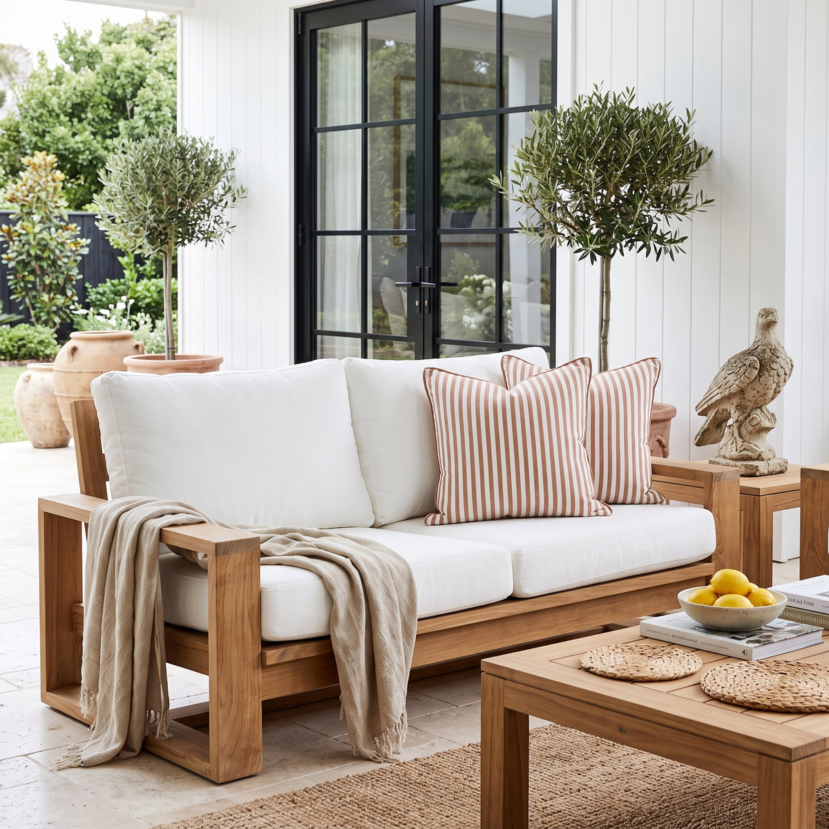 A wooden outdoor sofa with thick white cushions and striped peach-and-beige throw pillows, styled on a patio with a beige throw blanket, matching coffee table, potted trees, and a white shiplap wall background.