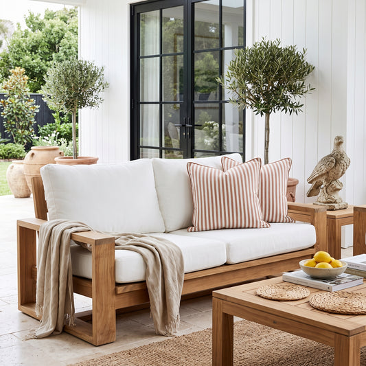 A wooden outdoor sofa with thick white cushions and striped peach-and-beige throw pillows, styled on a patio with a beige throw blanket, matching coffee table, potted trees, and a white shiplap wall background.