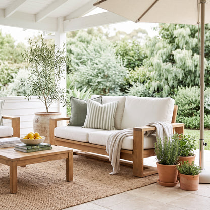 A wooden outdoor loveseat with off-white cushions and striped pillows, styled on a covered patio with a matching coffee table, potted plants, and a beige umbrella.