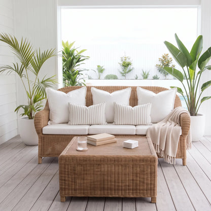 A wicker outdoor sofa set with white and cream cushions, styled with a matching wicker coffee table, a beige throw blanket, and potted plants in a bright, modern patio setting.